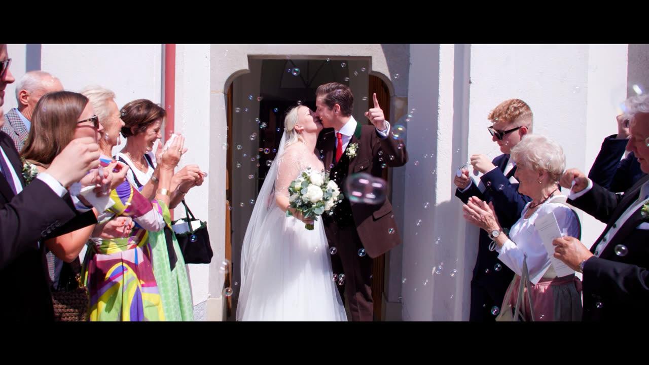 Brautpaar küsst sich im Kirchenportal, während die Hochzeitsgäste Seifenblasen pusten – Standbild aus dem Hochzeitsvideo von Eva-Maria und Hans in Oberösterreich