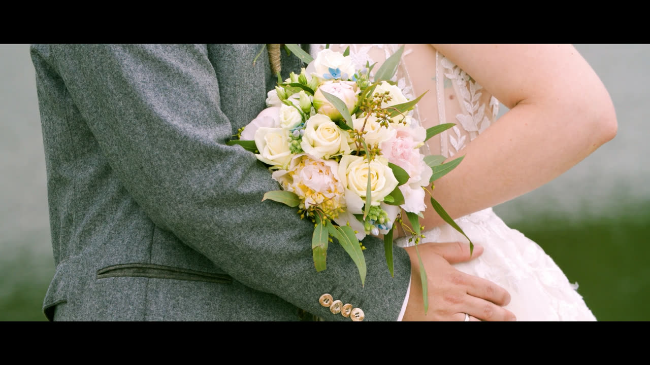 Nahaufnahme des Brautstraußes aus cremefarbenen Rosen und Eukalyptus, die Braut in Spitze an den Arm des Bräutigams im grauen Tweedsakko gelehnt – Standbild aus dem Hochzeitsvideo von Melissa und Marvin in der Steiermark